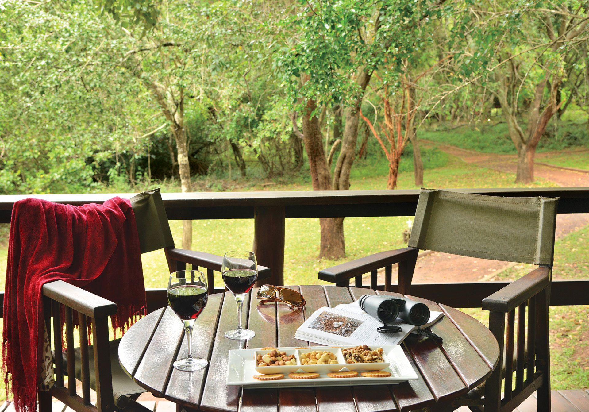 A wooden deck with a table set for a meal, glasses of red wine, and a plate of nuts and biscuits.
