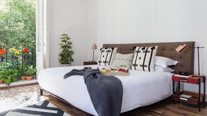 Uma cama bem arrumada com um cobertor cinzento, uma mesinha de cabeceira com abajur e uma pequena planta perto da janela.