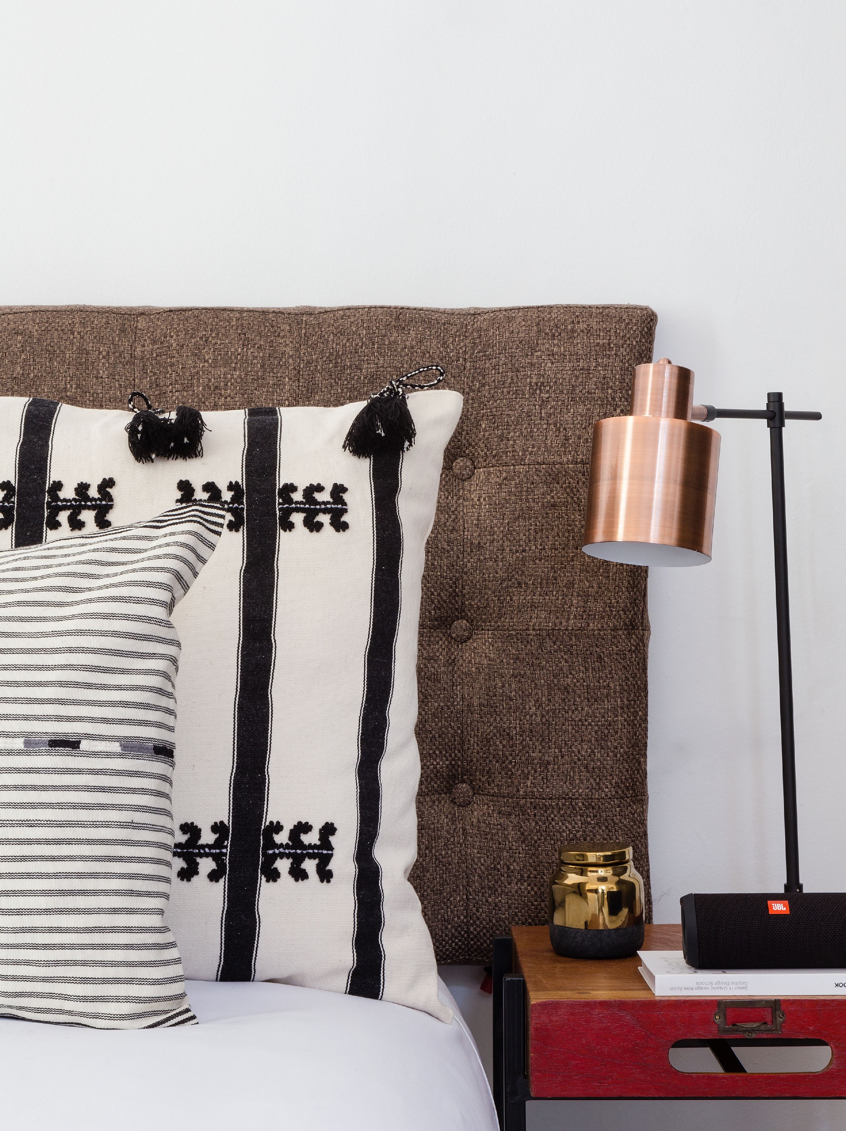 Un lit avec des oreillers à rayures, une lampe en cuivre, une petite statue dorée et une valise rouge posées sur une table de chevet en bois.