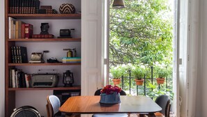 Uma sala de jantar com mesa e cadeiras de madeira, uma estante com diversos livros e objetos decorativos, e vista para o verde através de uma porta aberta.