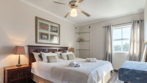 Un dormitorio ordenado con cama, mesitas de noche, ventilador de techo y ventana con cortinas.