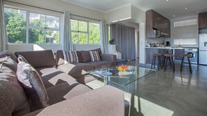 Una sala de estar moderna con una mesa de centro de vidrio, un sofá y una zona de cocina con taburetes de bar.