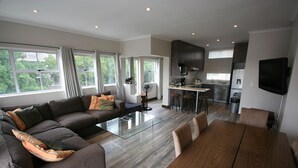 Una sala moderna con un amplio sofá seccional, una mesa de centro de vidrio y una mesa de comedor de madera. Cuenta con una zona de cocina con armarios oscuros y un televisor de pantalla plana montado en la pared.