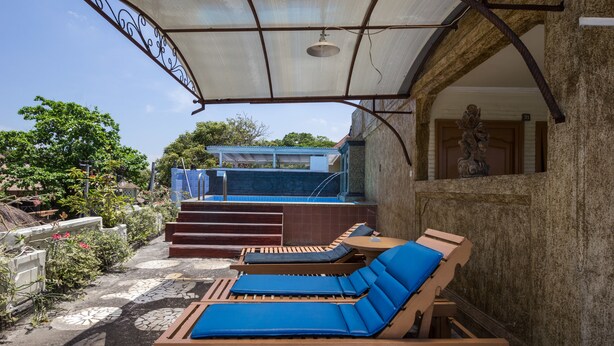 A covered outdoor seating area with two lounge chairs and a small table, a staircase leading to a blue-roofed building, and a statue in the background. - Reddoorz @ Cemara Beach Sunrise (Denpasar)