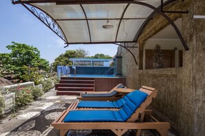 A covered outdoor seating area with two lounge chairs and a small table, a staircase leading to a blue-roofed building, and a statue in the background. - Reddoorz @ Cemara Beach Sunrise (Denpasar)