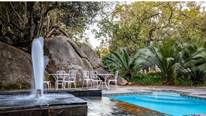 Une piscine agrémentée d’une cascade, entourée d’un espace détente avec des chaises et une table, le tout mis en valeur par un décor de gros rochers et d’une végétation luxuriante.