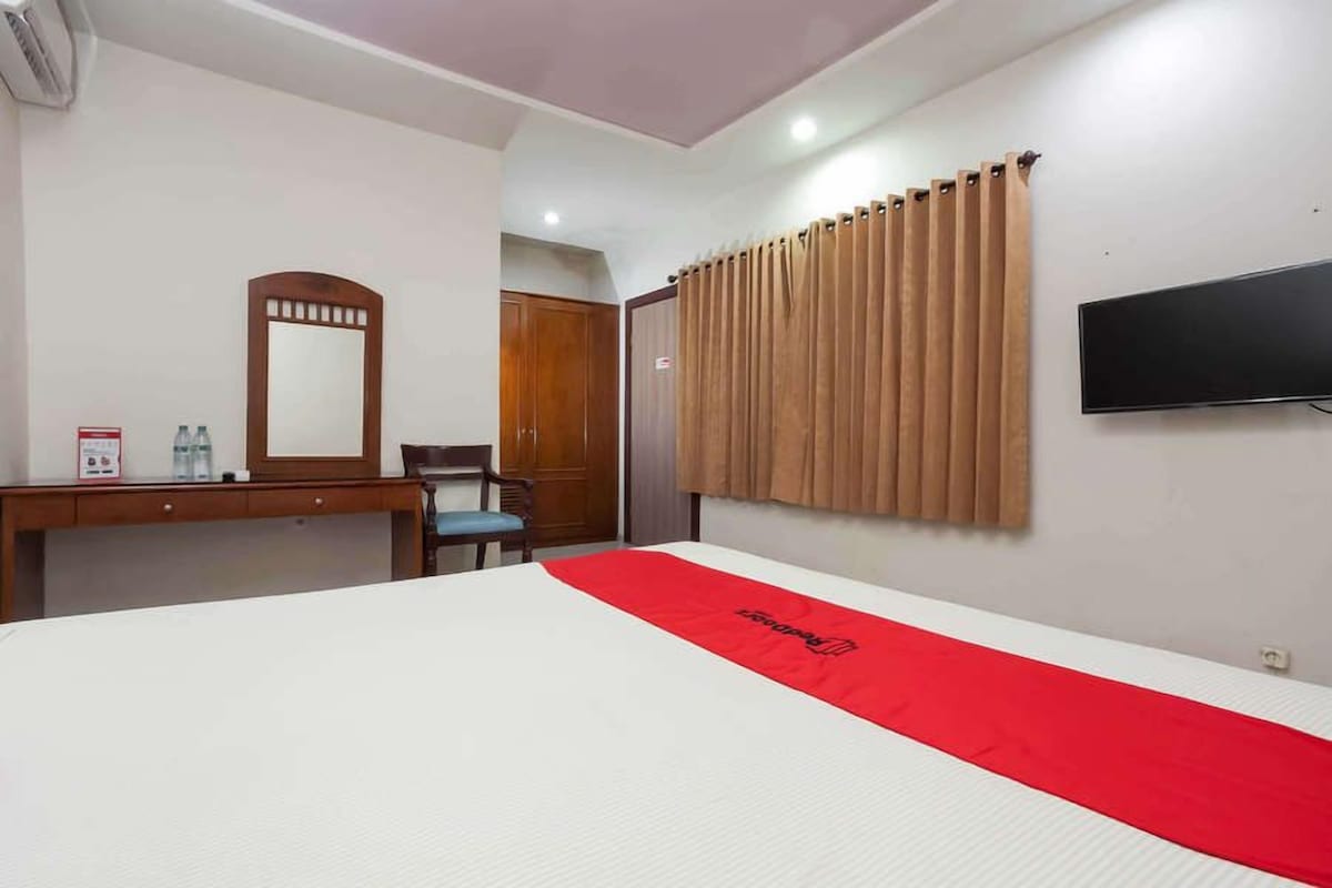 Habitación de hotel con una cama grande, un escritorio de madera, una silla, un espejo, un televisor y una ventana con cortinas.