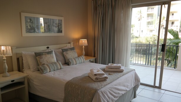 A hotel room with a large bed, two bedside tables with lamps, a view of the balcony, and a painting on the wall. - Bougain Villas Apartments (Cape Town)