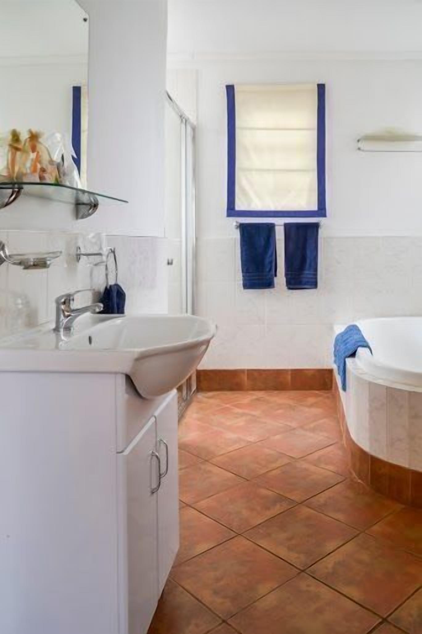 Een badkamer met een wit wastafel, een spiegel, een glazen plankje, een raam met een blauwe handdoek, en een bad.