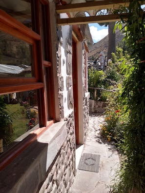 A stone-walled corridor with a window and a door, leading to a garden with various plants and flowers. - Samana Wasi (Yanque)