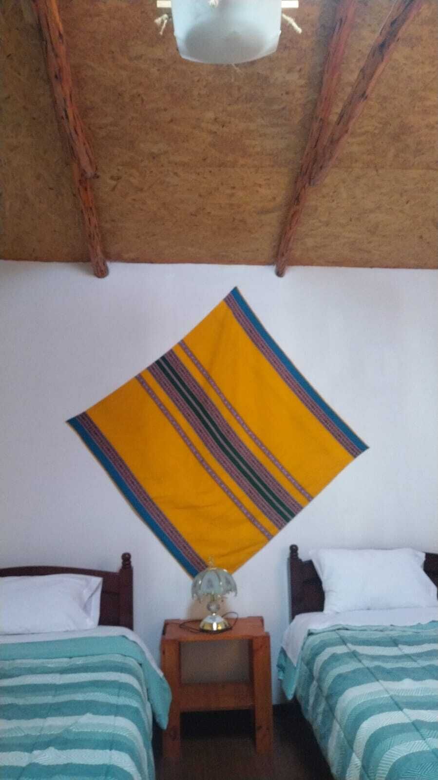 A room with two beds, a wooden nightstand with a lamp, and a hanging yellow and blue striped blanket.