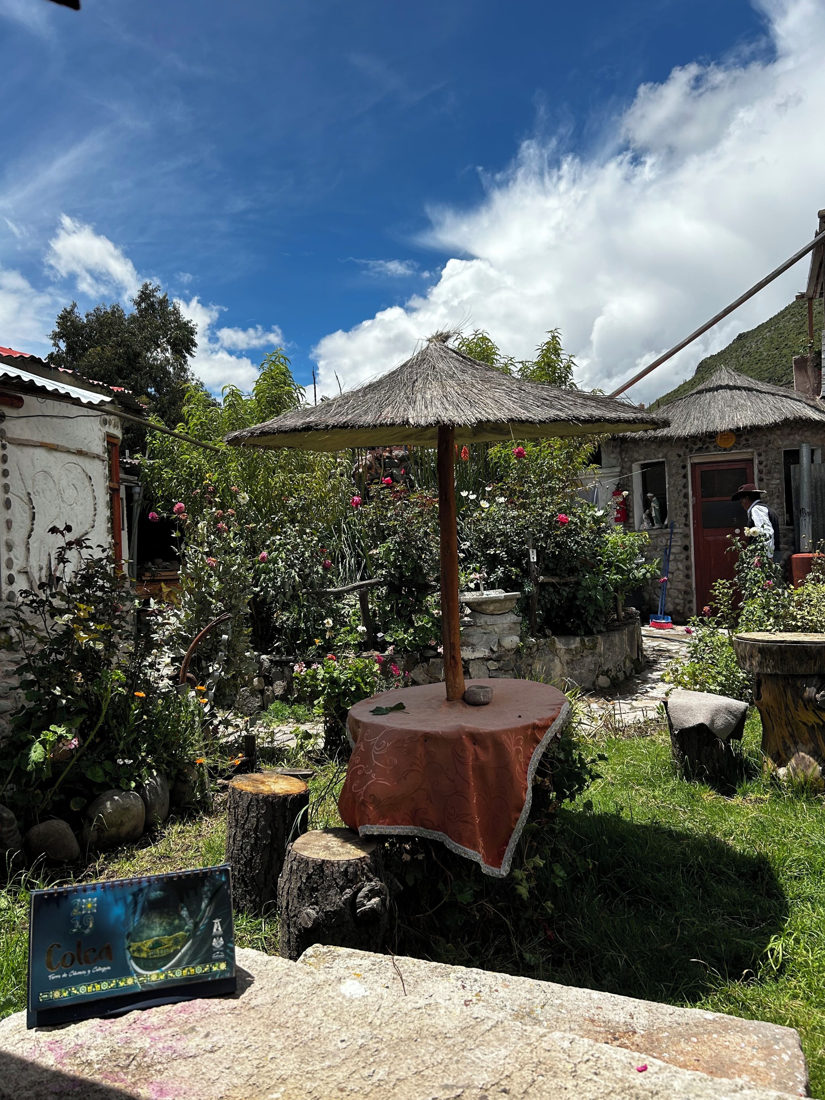 Un entorno rústico al aire libre con una mesa de techo de paja, un banco de piedra y un letrero.