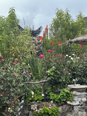 Un jardín con diversas plantas y flores, un muro de piedra y una estructura rústica con techo de paja.