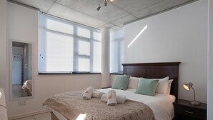 A hotel room with a large bed, a nightstand, a mirror, and a window with blinds. - Hillside Heights 503 by CTHA (Cape Town)
