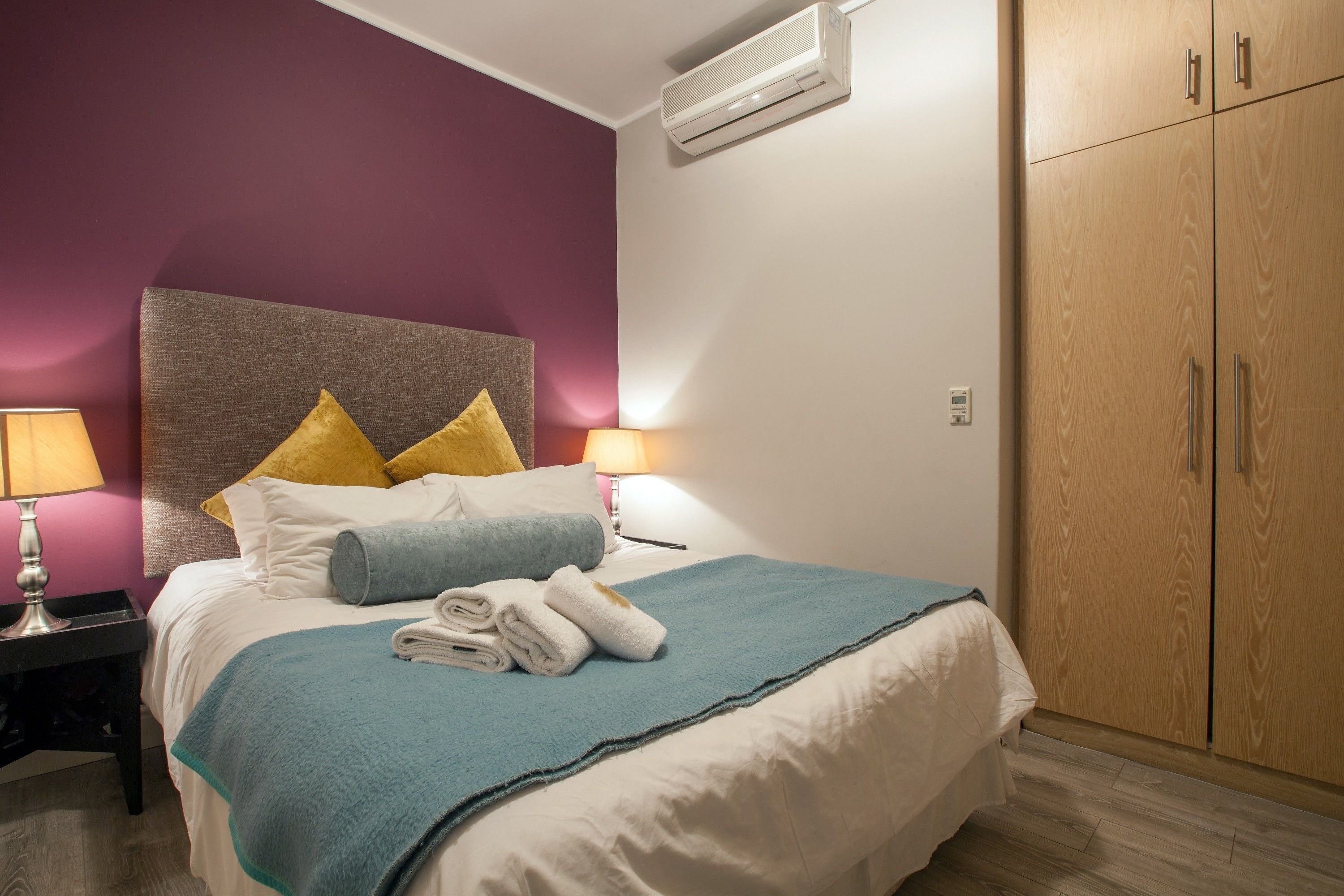 A neatly made bed with a blue and white duvet, two pillows, and folded towels. There is a wooden wardrobe with two doors and a nightstand with a lamp.