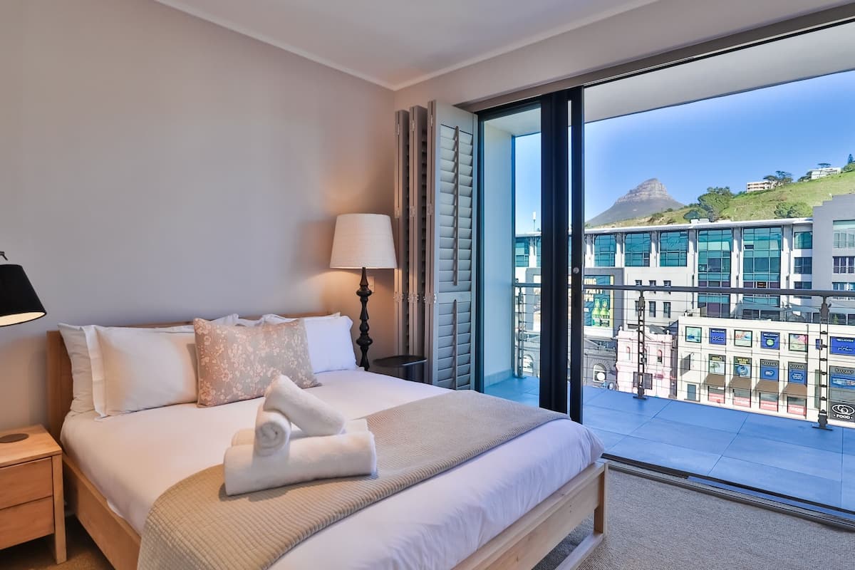 Habitación de hotel con una cama grande, una mesita de noche, una lámpara y vistas a edificios y una colina.