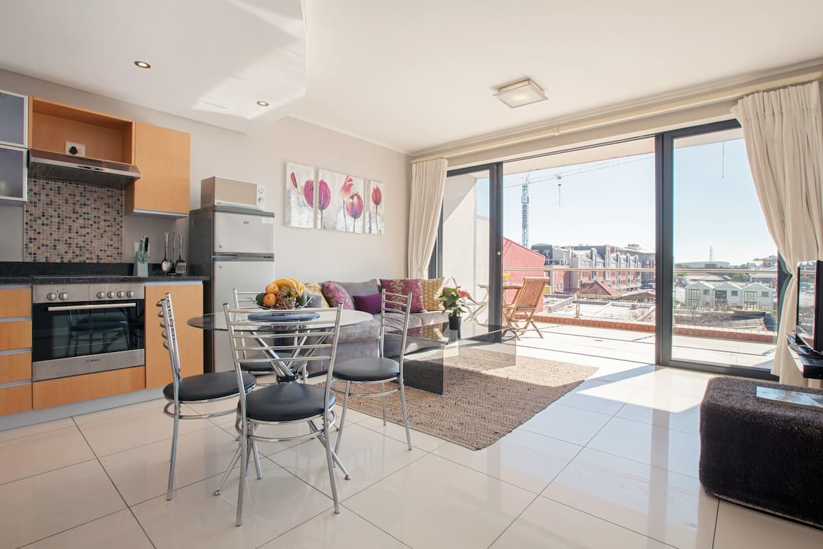 Una cocina moderna con electrodomésticos de acero inoxidable, un comedor con mesa y sillas de vidrio, y un amplio ventanal corredero que da a un balcón con vistas a la ciudad.