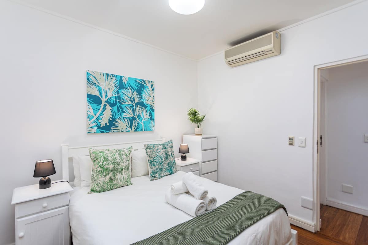 Un dormitorio ordenado con cama, mesitas de noche, aire acondicionado de pared y una puerta que da a otra habitación.