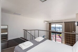 A hotel room with a bed, a small seating area, a window with a view, a balcony, and a staircase.