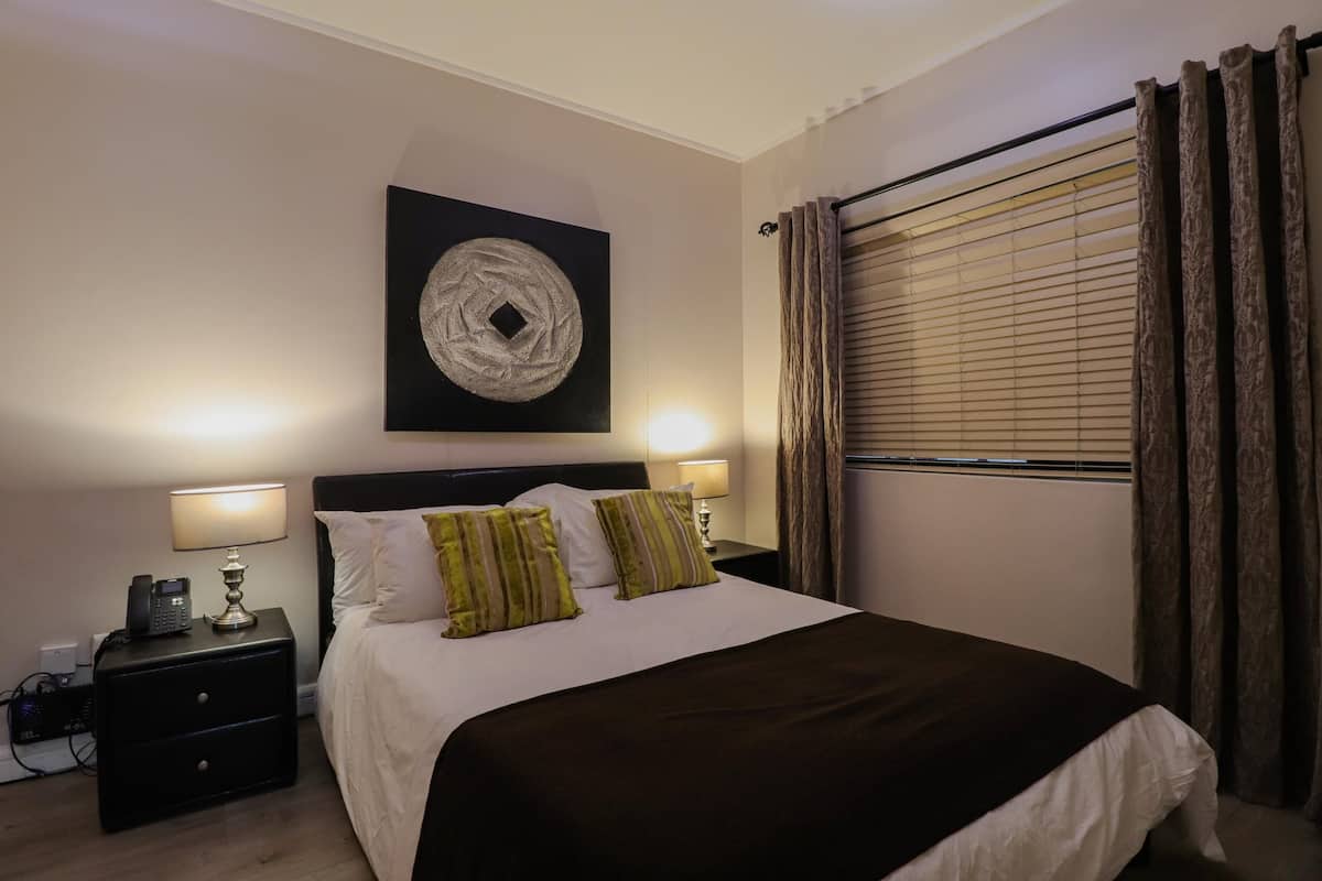 Una cama bien hecha con un edredón oscuro y dos almohadas, una mesita de noche con una lámpara, una obra de arte en la pared y una ventana con cortinas.