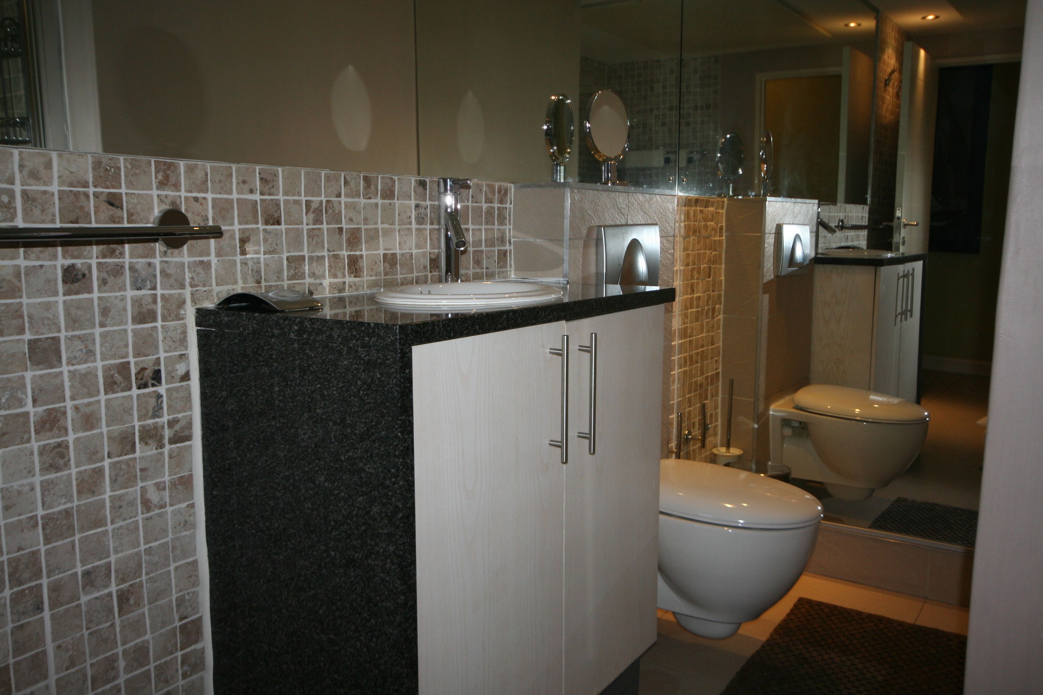 A modern bathroom with a granite countertop, white cabinets, a white toilet, and a glass shower enclosure.