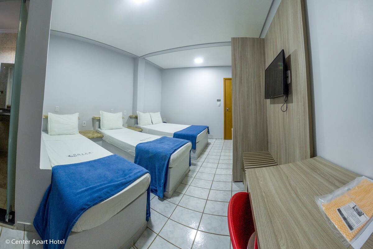 A hotel room with three single beds, a wooden desk, a red chair, and a television on the wall.