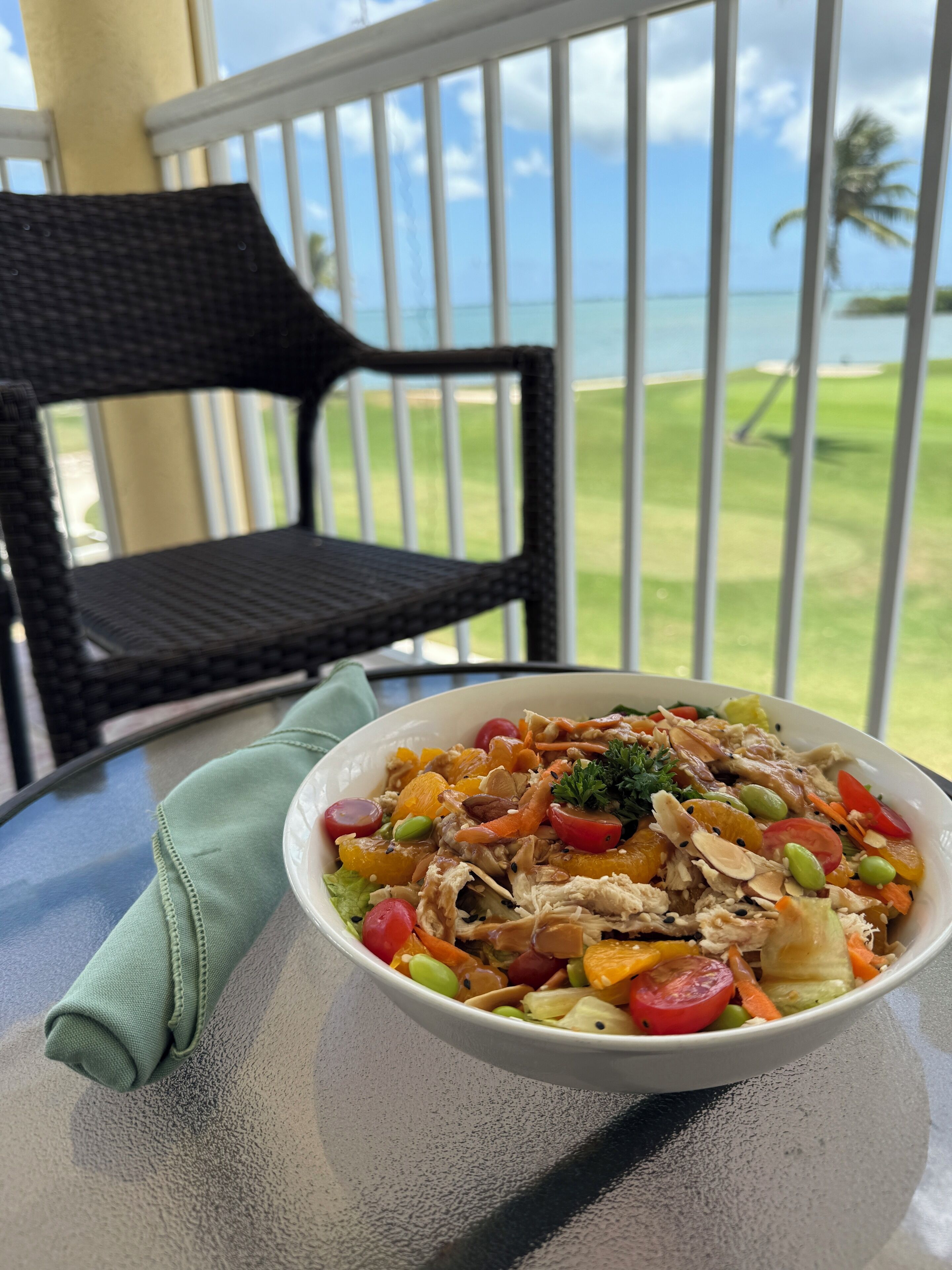 A bowl of salad on a table with a view of the ocean and a chair in the background.