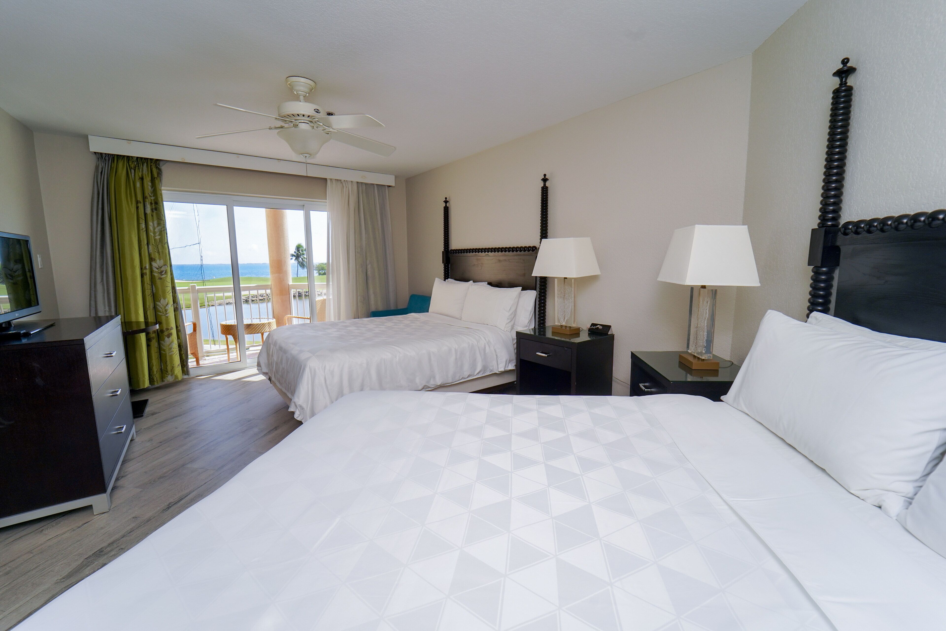 Une chambre d’hôtel avec deux lits, un ventilateur de plafond, une télévision et un balcon avec vue sur l’océan.
