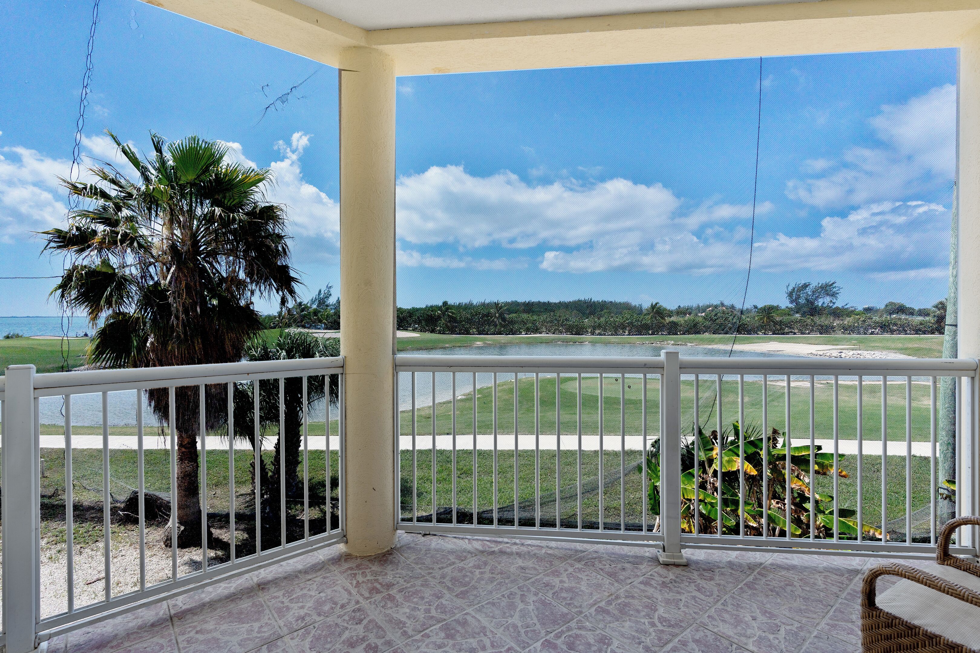 A covered balcony with a view of a golf course, palm tree, and a wicker chair.