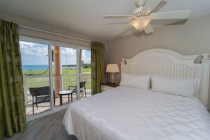 Habitación de hotel con cama, ventilador de techo, mesita de noche con lámpara, una silla y vista al océano a través de amplias ventanas.