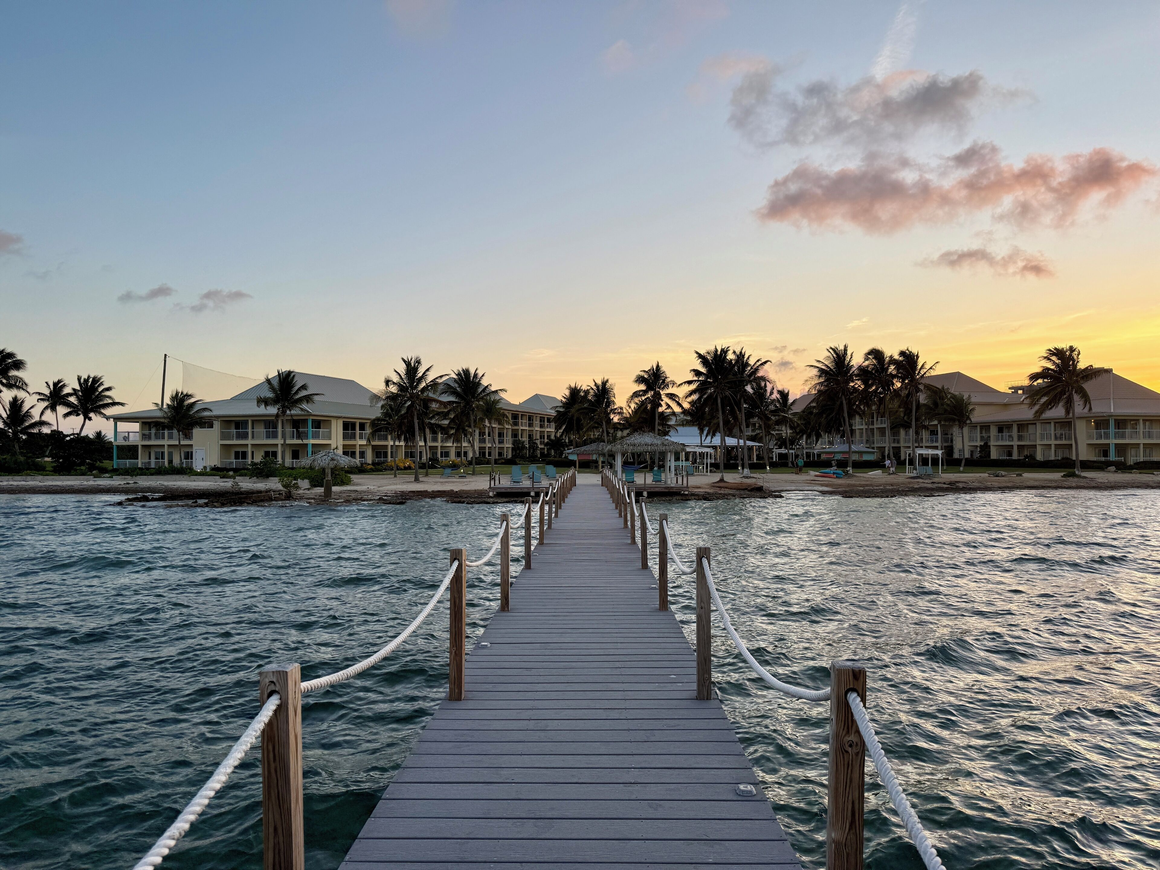 A wooden pier leading to a beachfront hotel with multiple balconies and palm trees.
