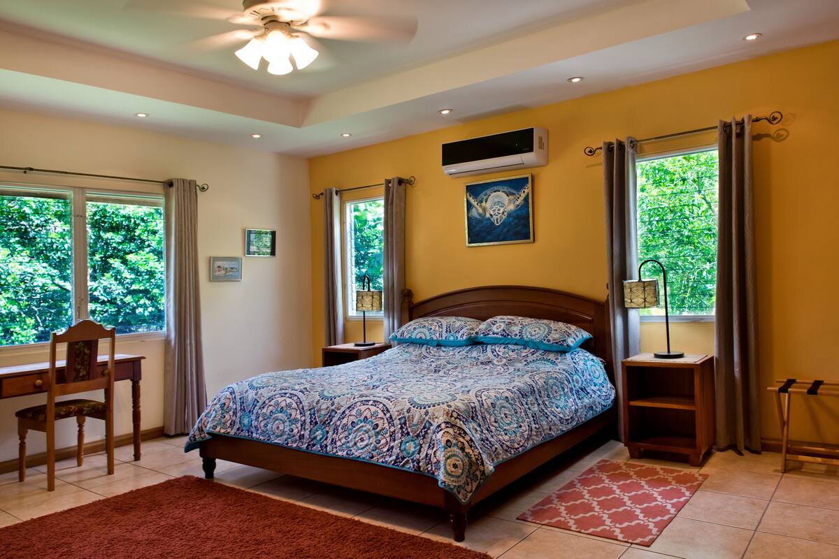 Ein Schlafzimmer mit einem Bett, einem Schreibtisch, einem Stuhl, einem Fenster mit Vorhängen, einer Klimaanlage und einem Deckenventilator.