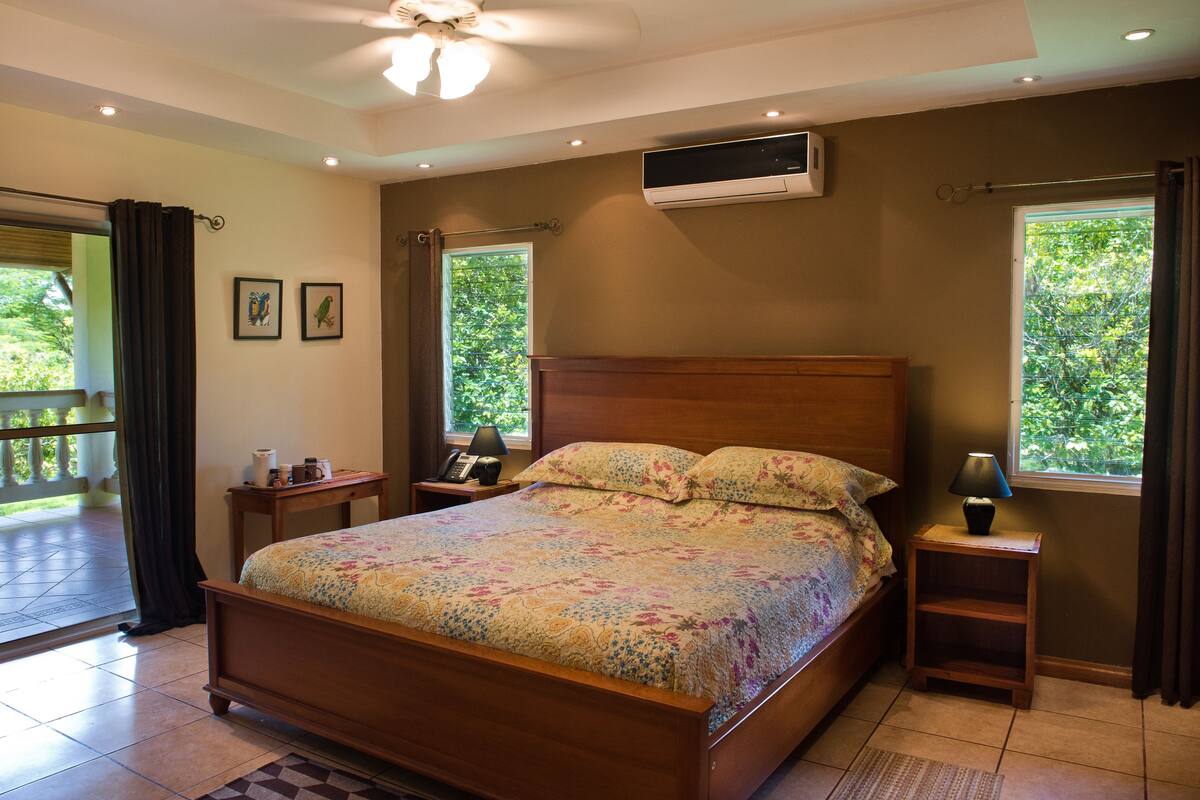 Ein Schlafzimmer mit einem hölzernen Bett, zwei Nachttischen, einem kleinen Tisch und einer Klimaanlage, sowie einem Balkon mit Blick auf Grünflächen.
