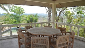 Un espacio de estar al aire libre con techo, con una mesa redonda y sillas de madera, con vistas a un paisaje exuberante con árboles y una cadena montañosa distante.