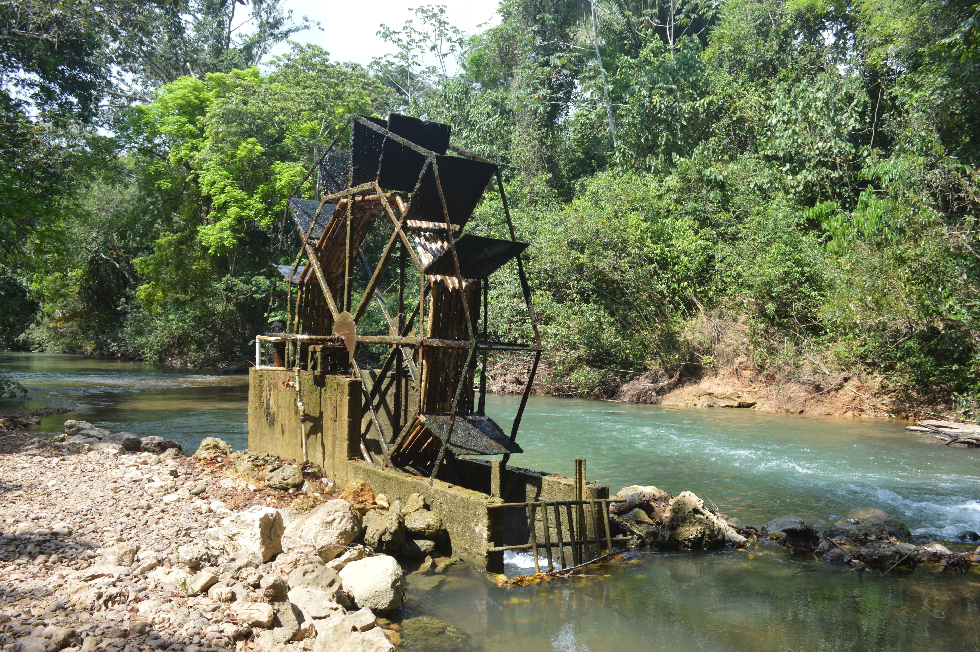 A rustic water wheel structure by a river, surrounded by dense greenery.