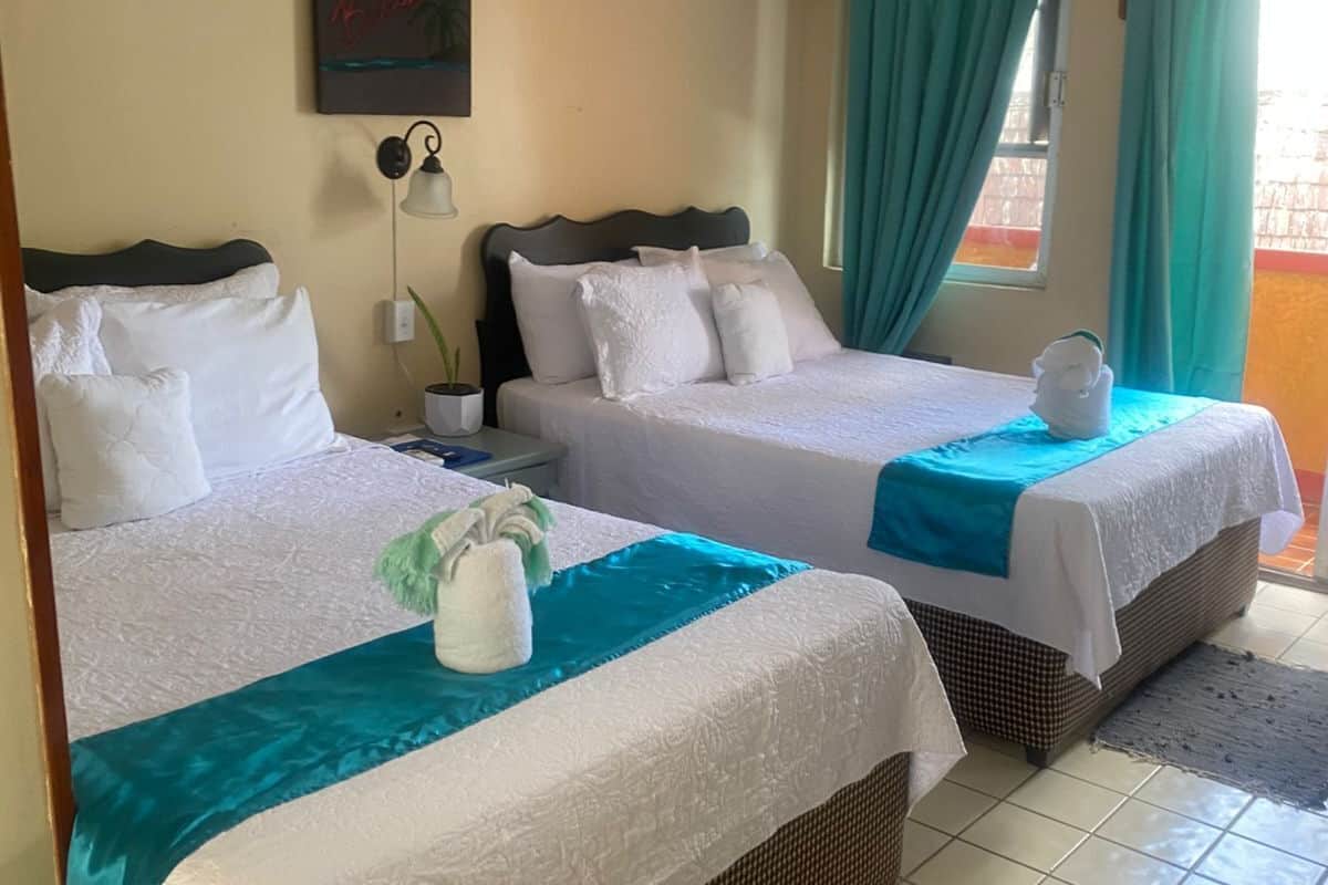 Habitación de hotel con dos camas, aire acondicionado de pared, mesita de noche con lámpara y ventana con cortinas.