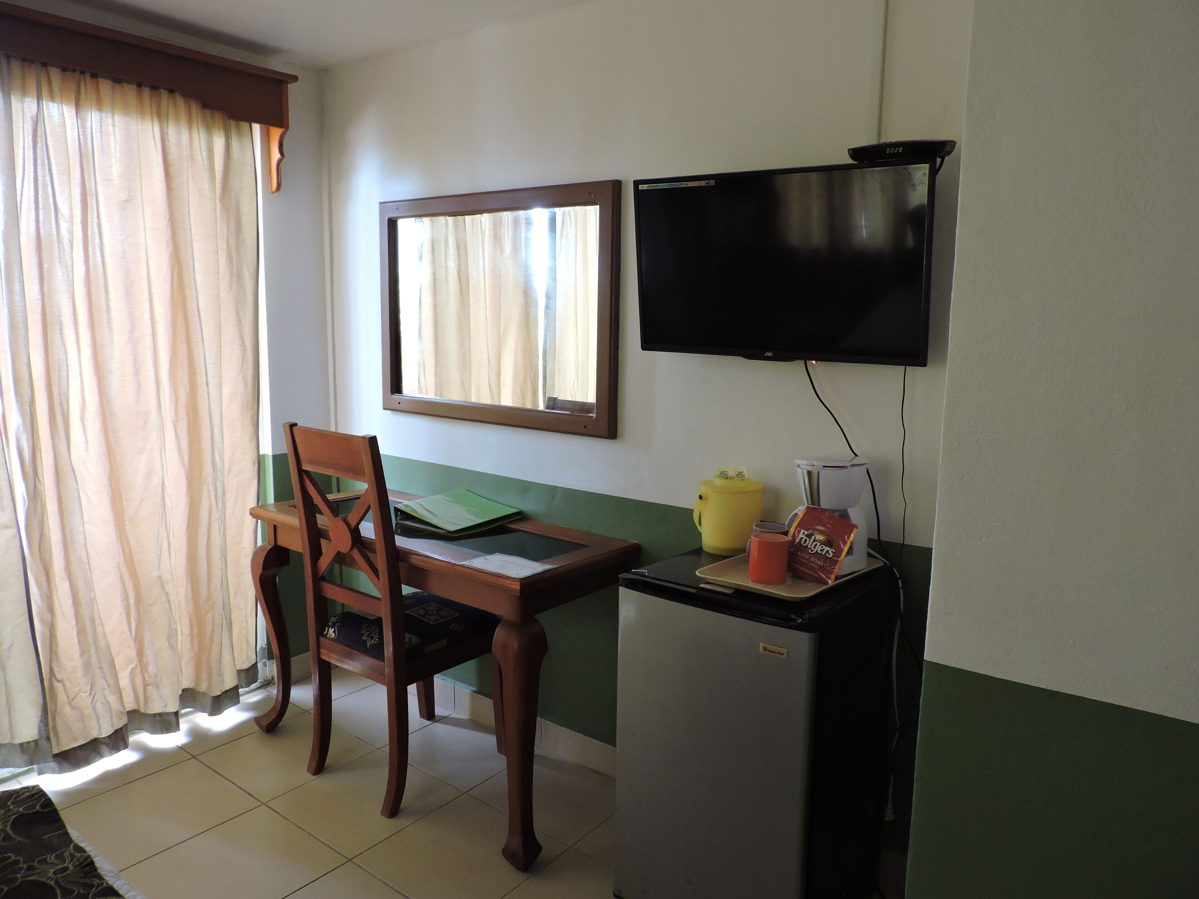 A room with a wooden desk, a chair, a television mounted on the wall, a refrigerator, and a window with curtains.