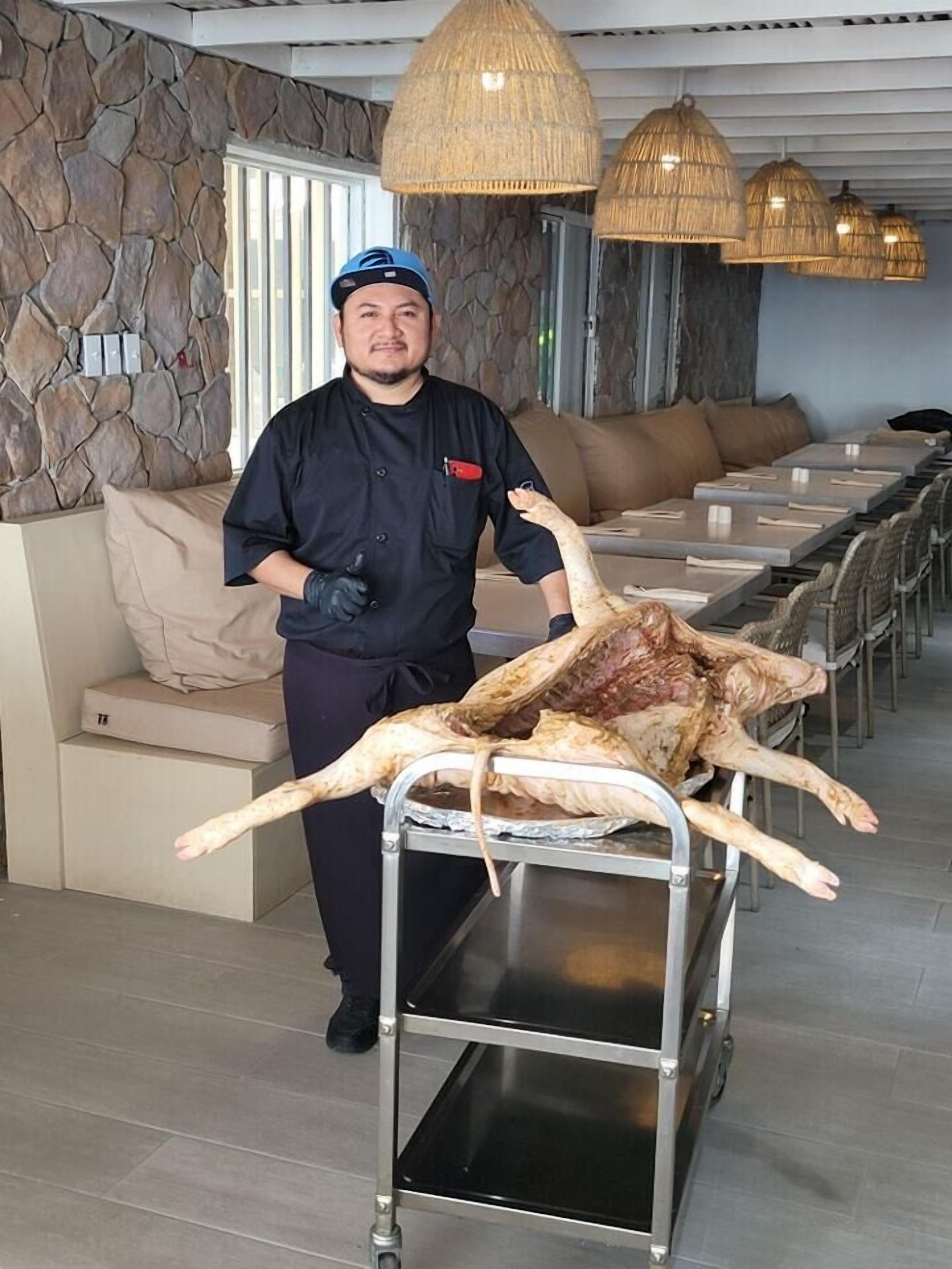 A person in a black uniform pushing a cart with a large animal carcass in a restaurant setting with stone walls and wicker ceiling lights.