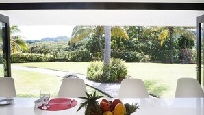 Una mesa de comedor con un frutero y un vaso de agua, con vistas a un jardín exuberante con palmeras y un sendero.