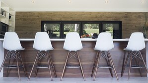 Una cocina moderna con un muro decorativo de madera, un ventanal grande y cuatro taburetes blancos.
