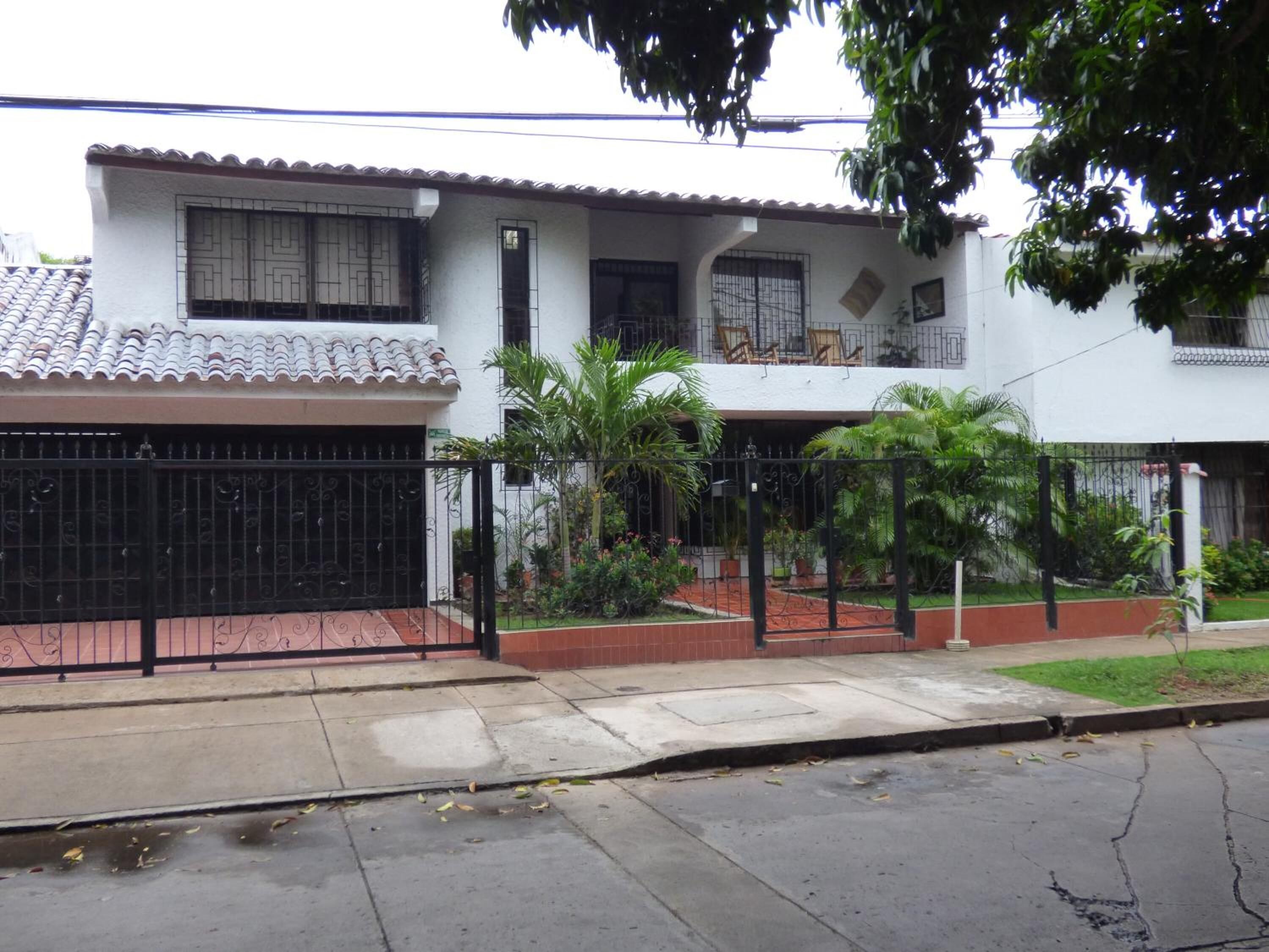 A two-story house with a tiled roof, a black metal gate, and a small garden with palm trees.