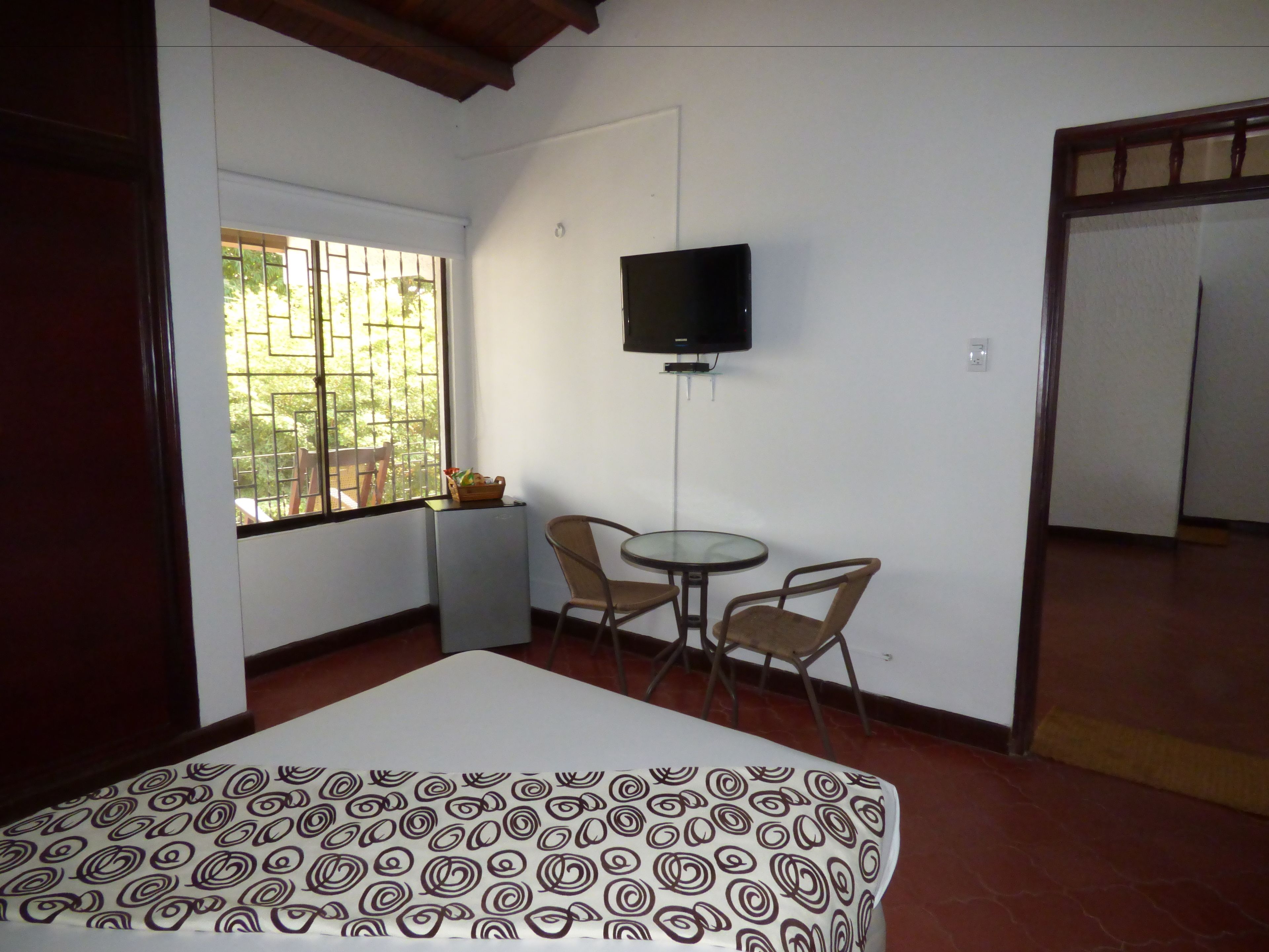 A room with a bed, a small table, two chairs, a TV mounted on the wall, and a window with bars.