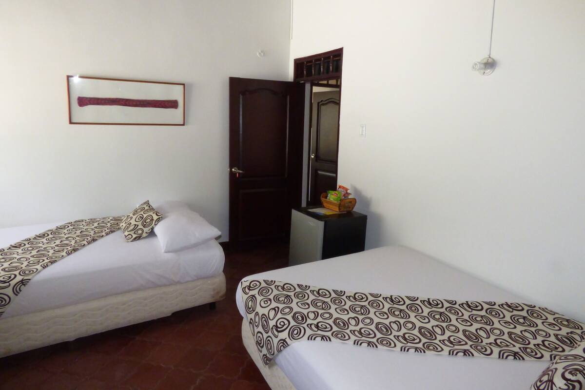 A hotel room with two single beds, a wooden door, a small table with snacks, and a framed picture on the wall.