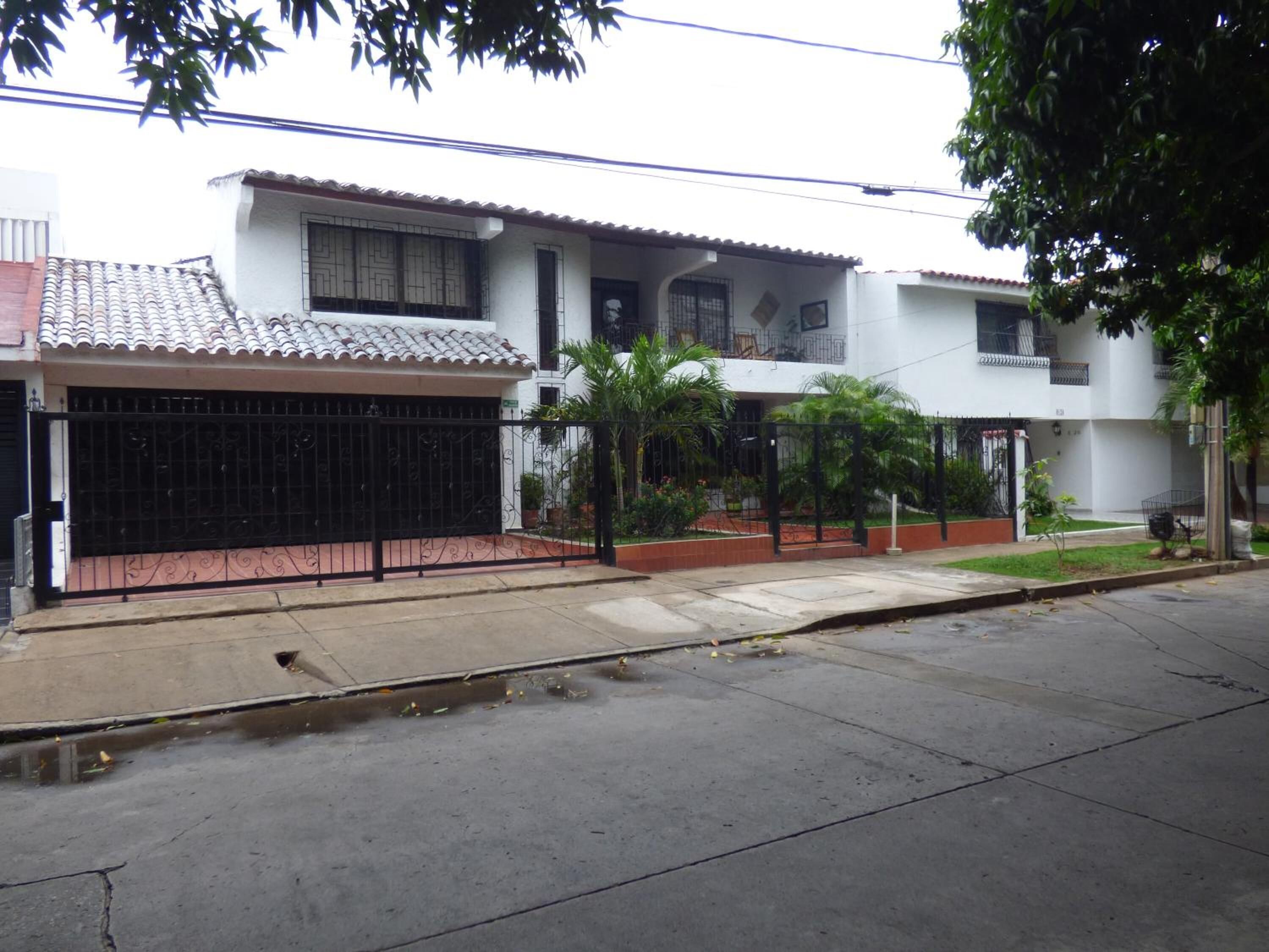 A two-story house with a tiled roof, a black metal gate, and a small garden with palm trees.