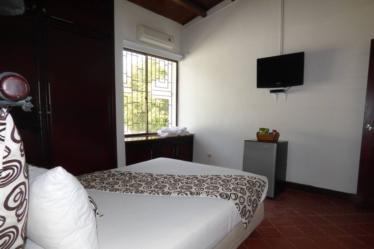 A bedroom with a bed, a TV mounted on the wall, a small cabinet, and a window with bars.