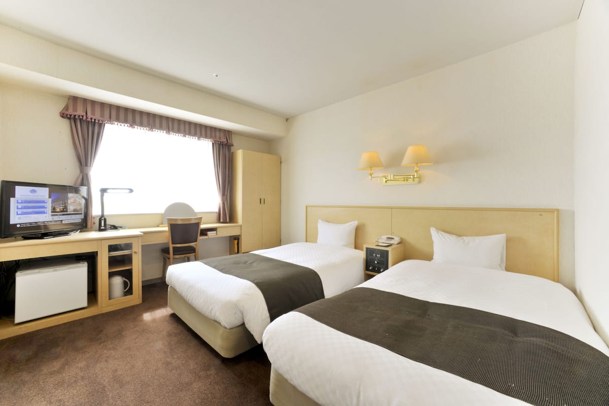Une chambre d’hôtel avec deux lits, un bureau avec un ordinateur, une chaise, un petit réfrigérateur et une fenêtre avec des rideaux.