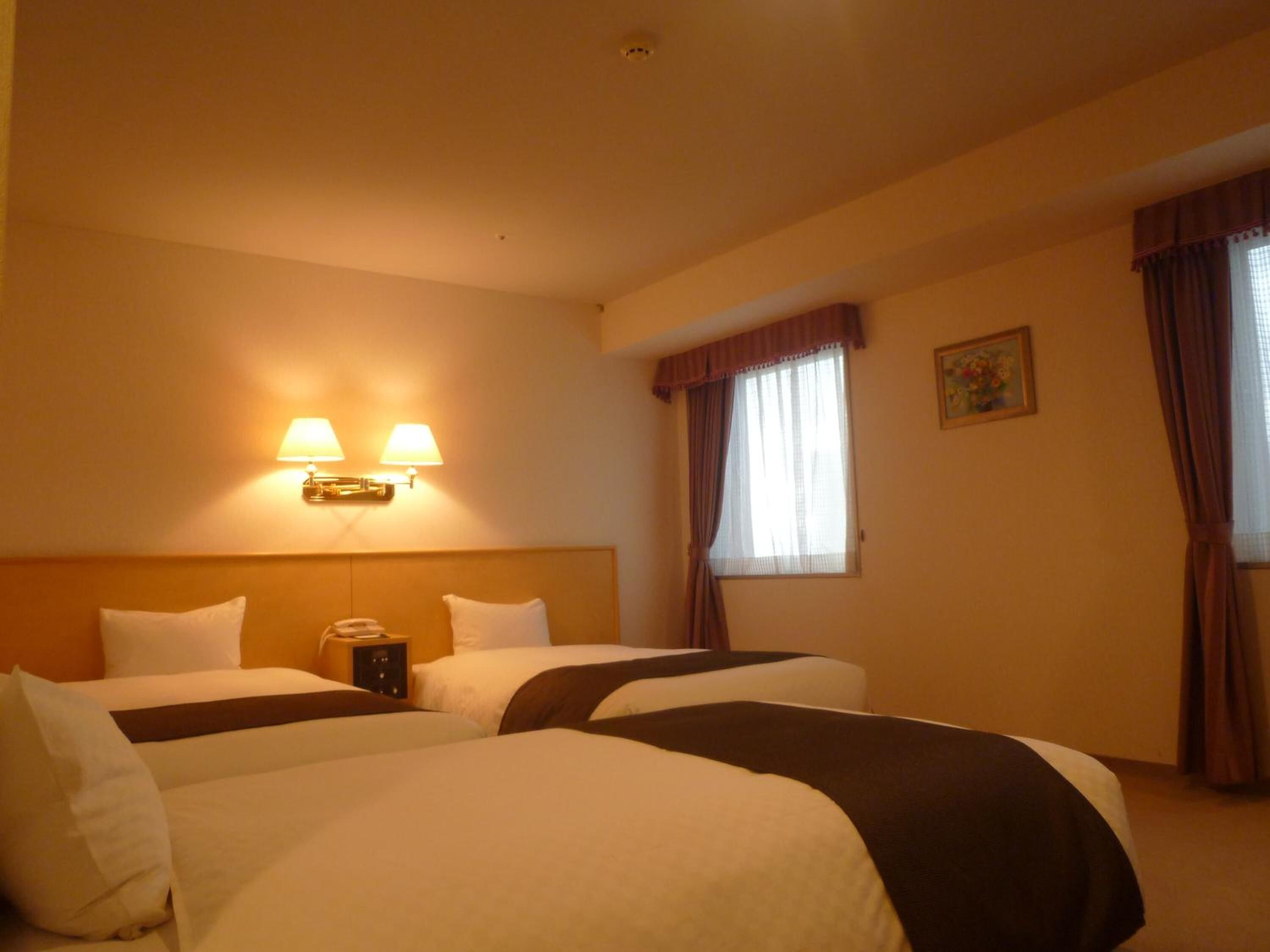 Une chambre d’hôtel avec deux lits, une table de chevet, une lampe, une fenêtre avec des rideaux et un tableau au mur.