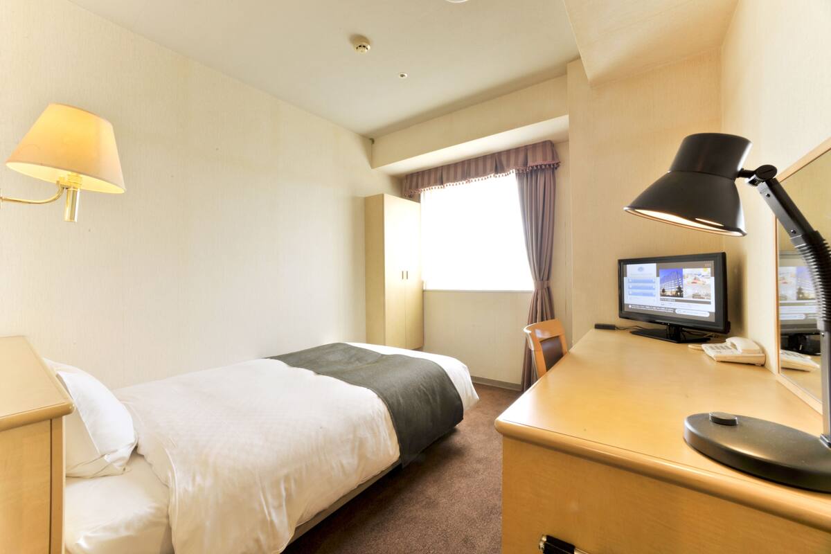 Une chambre d’hôtel avec un lit, un bureau avec un ordinateur, une lampe et une fenêtre avec des rideaux.