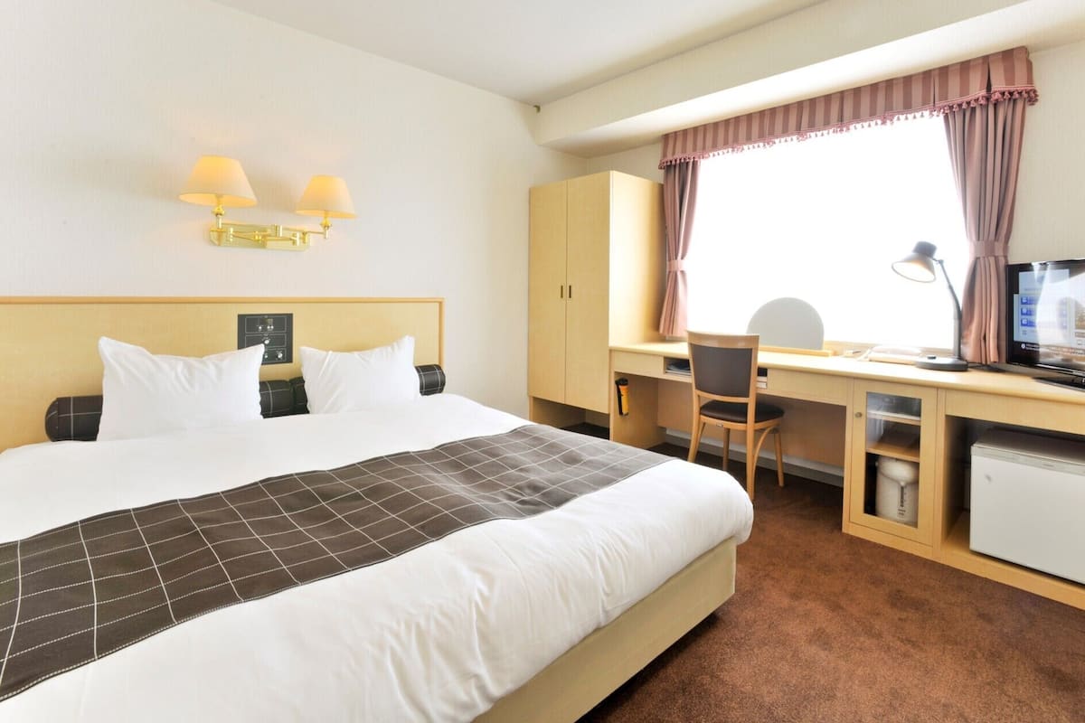 Quarto de hotel com cama, escrivaninha, cadeira, televisão e um pequeno frigorífico.