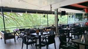 An outdoor seating area with tables and chairs, a view of greenery, and a glass wall with a bar area. - Hotel Gangaaddara (Kandy)